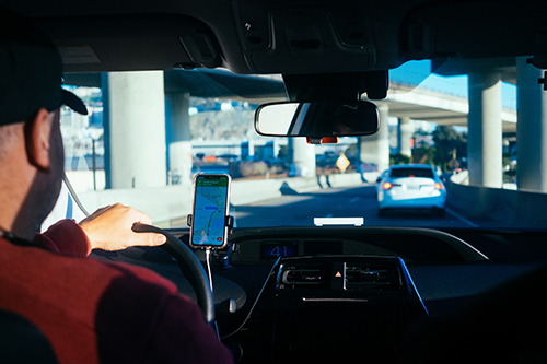 Photo taken from the backseat of a Lyft vehicle. Driver is seen in profile to the left of the picture. Phone is mounted on the dash and displays a maps app that is tracking the route to the passenger&#039;s destination. Photo by Paul Hanaoka on Unsplash.