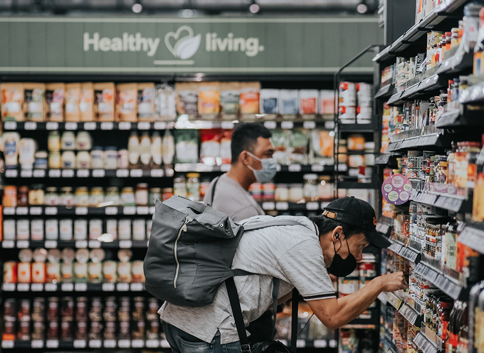 A man is seen grocery shopping during the pandemic, looking at canned food while wearing a mask.