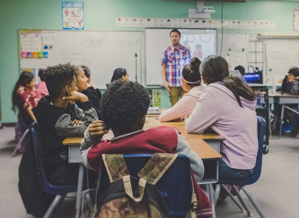 A photo of children listening to teacher in class, taken from a child&#039;s perspective.