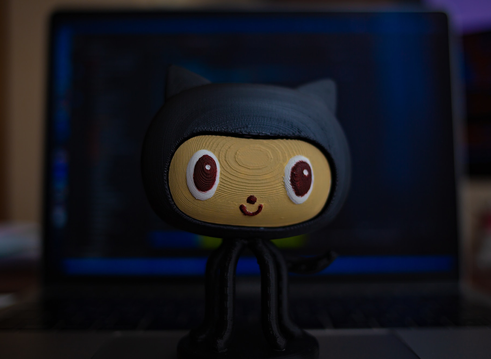 A small figurine of an oktokata in the center, in the background a laptop with an open code editor and a terminal.